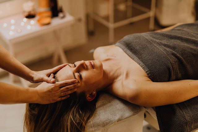 A woman lies on a treatment couch with her eyes closed while a therapist cradles her face during a facial, warm candlelit clinic room